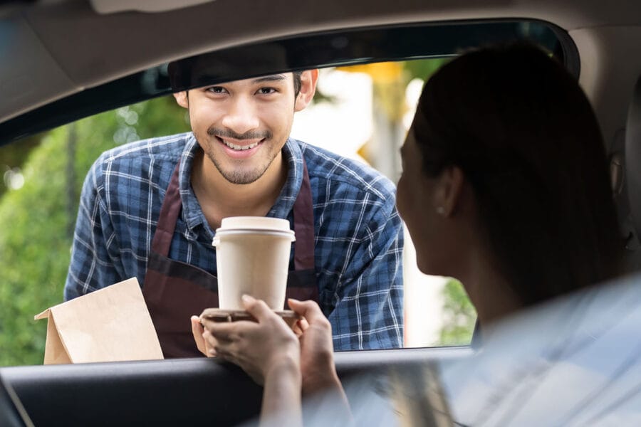 Man working at coffee drive thru hands a to-go cup of hot coffee to woman in passenger seat through the car window